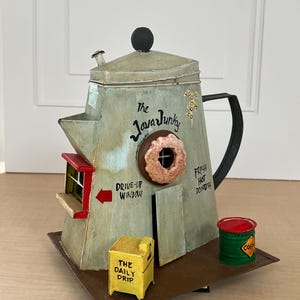 May include: A white ceramic mug with a black handle and the text "But First, Coffee" in a bold, black font. The mug is set against a blurred background, suggesting a cozy setting.