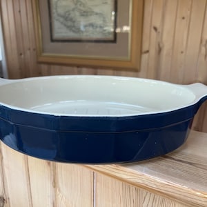 May include: A navy blue oval baking dish with a cream-colored interior. The dish has two handles and is made of a glossy material. This kitchenware is suitable for baking and serving.