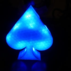 Op de afbeelding: Een schoppenvormig object met een doorschijnend oppervlak dat oplicht met een felblauw licht. De structuur heeft een zichtbare rasterachtige textuur over het oppervlak, waardoor een geometrisch patroon ontstaat dat van binnenuit blauw wordt verlicht.