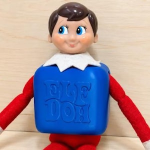 May include: A doll with brown hair, blue eyes, and rosy cheeks wearing a white collar. The doll has a blue plastic torso with the embossed text ELF DOH on the front, and long red fabric arms with white cuffs extending from the sides.