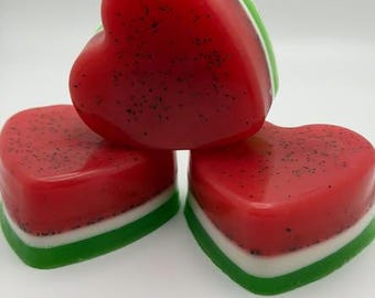 May include: Three heart-shaped bars of soap featuring a layered design that mimics a watermelon. Each bar has a translucent red top layer with small black specks, a thin white middle layer, and a solid green bottom layer.