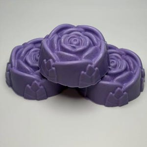 May include: Three lavender-colored bars of soap, each molded into the shape of a blooming rose. The soap bars feature detailed petal textures on the top surface and a smooth, rounded base with small, decorative leaf-like accents along the sides.