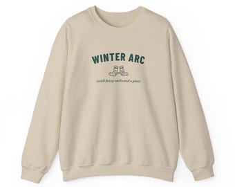 以下が含まれることがあります： 胸の中央にダークグリーンの文字で「WINTER ARC」とプリントされたベージュのクルーネックトレーナー。文字の下には靴下2足の小さなグラフィックと、ダークブラウンのフォントで括弧書きの「(with fuzzy socks and a plan)」というフレーズが配置されています。