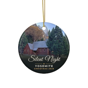 Può includere: Una decorazione natalizia rotonda in ceramica con la fotografia di una cappella in legno rosso a Yosemite circondata da alberi. Il testo recita Silent Night, YOSEMITE e CHRISTMAS 2026 in lettere bianche e dorate. Appesa a un filo dorato.