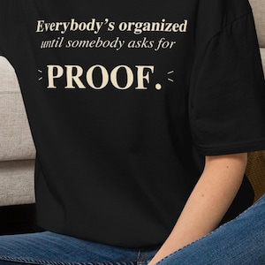 May include: A black short-sleeved t-shirt featuring the cream-colored text "Everybody's organized until somebody asks for PROOF." The word PROOF is centered and emphasized with small decorative lines on either side of the word.