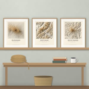 May include: Three framed prints featuring topographic maps of Kilimanjaro, Mont Blanc, and Mount Rainier. Each print displays the mountain name and coordinates in black text on a cream background, housed in light wood frames.