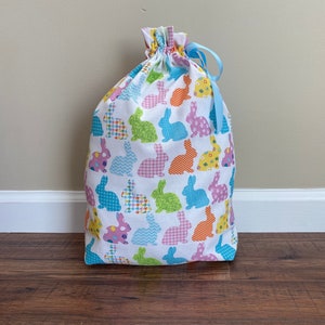 May include: A white drawstring bag with a colorful bunny pattern. The bunnies are in various colors and patterns, including blue, green, pink, orange, and yellow. A light blue ribbon is tied at the top of the bag.
