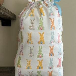A large reusable gift bag featuring bunnies holding a decorated egg printed on white with a light blue ribbon from the Easter and springtime collection at Reese and Co Shop