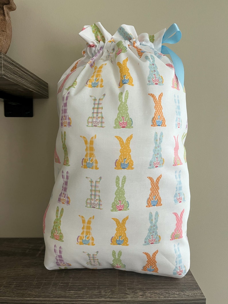 A large reusable gift bag featuring bunnies holding a decorated egg printed on white with a light blue ribbon from the Easter and springtime collection at Reese and Co Shop