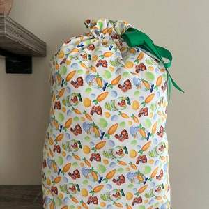 A large reusable gift bag featuring chocolate bunnies, carrots and eggs scattered on a white background with a dark green ribbon from the Easter and springtime collection at Reese and Co Shop