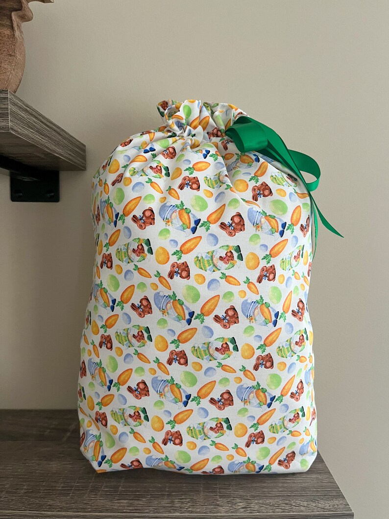 A large reusable gift bag featuring chocolate bunnies, carrots and eggs scattered on a white background with a dark green ribbon from the Easter and springtime collection at Reese and Co Shop