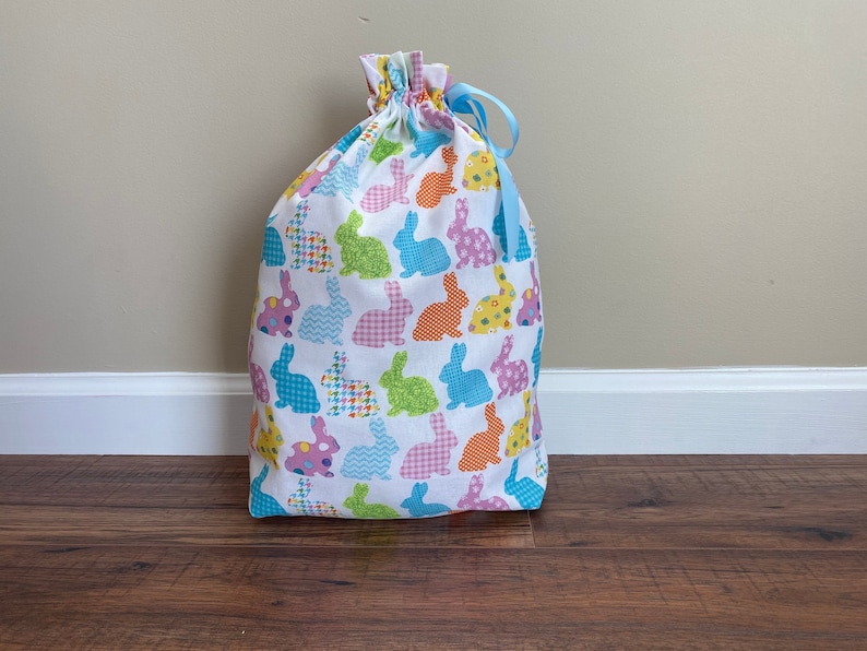 May include: A white drawstring bag with a colorful bunny pattern. The bunnies are in various colors and patterns, including blue, green, pink, orange, and yellow. A light blue ribbon is tied at the top of the bag.