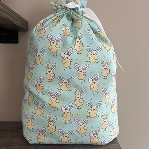 A large reusable gift bag featuring light yellow chicks with bunny ears scattered on a light blue fabric with a white ribbon from the Easter and springtime collection at Reese and Co Shop