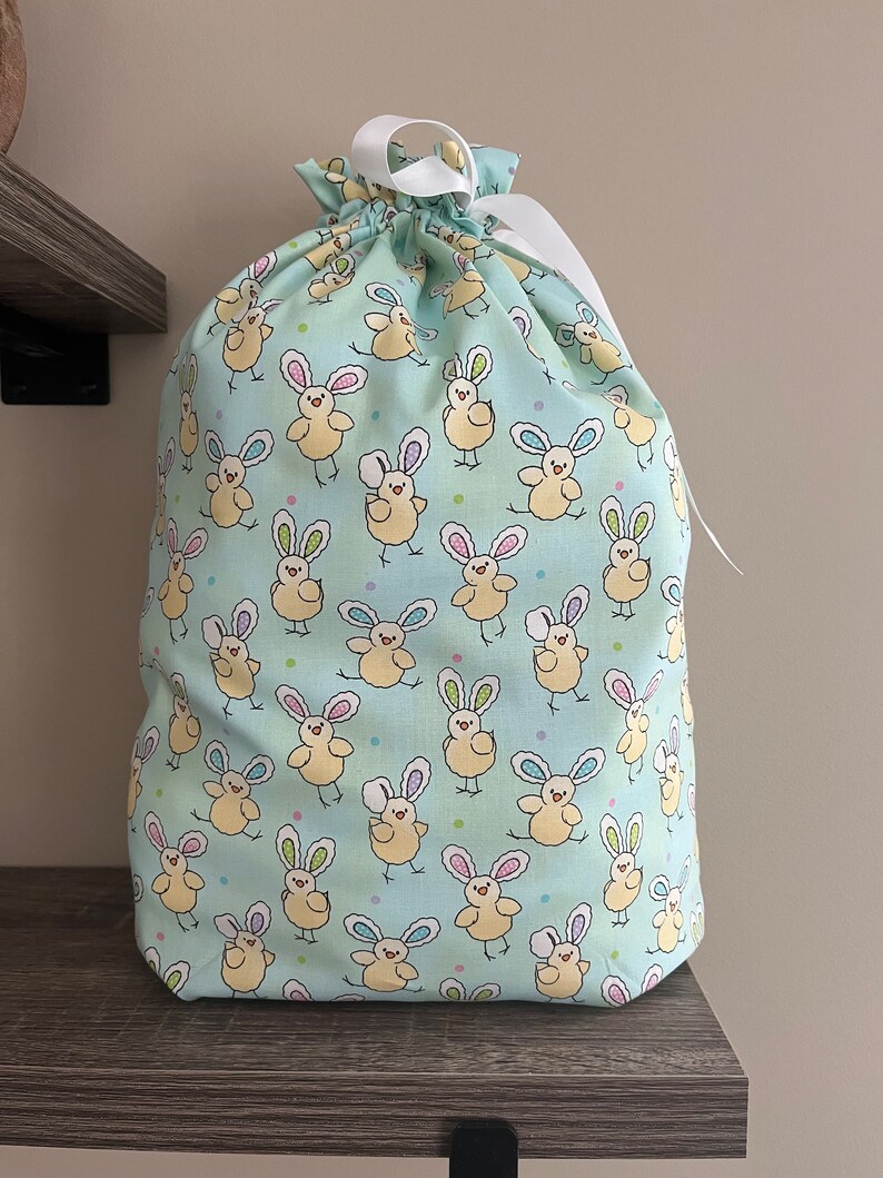 A large reusable gift bag featuring light yellow chicks with bunny ears scattered on a light blue fabric with a white ribbon from the Easter and springtime collection at Reese and Co Shop