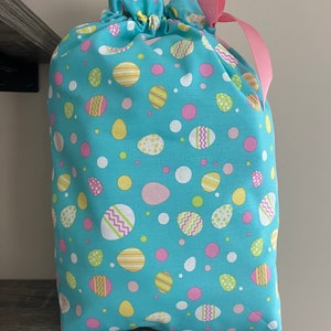 A large reusable gift bag featuring decorated pastel Easter eggs in various sizes scattered on a teal blue fabric with a pink ribbon from the Easter and springtime collection at Reese and Co Shop