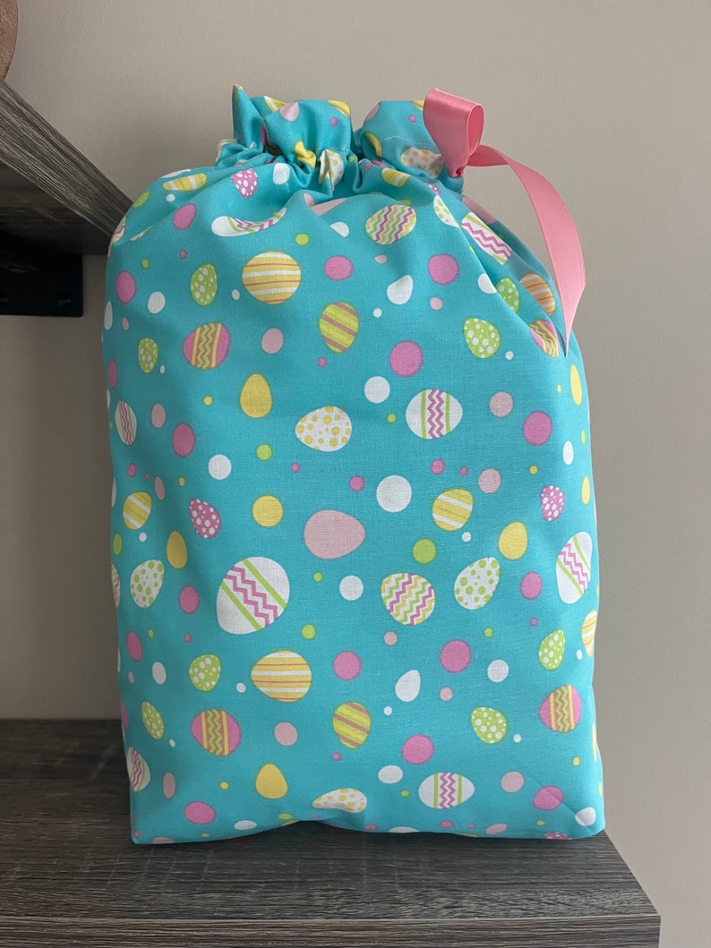 A large reusable gift bag featuring decorated pastel Easter eggs in various sizes scattered on a teal blue fabric with a pink ribbon from the Easter and springtime collection at Reese and Co Shop
