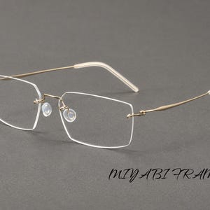 Puede incluir: Gafas sin montura con lentes rectangulares, patillas de metal fino en tono dorado y almohadillas nasales transparentes. Los extremos de las patillas están cubiertos de plástico blanco. El texto MIYABI FRAME aparece en negro abajo a la derecha.