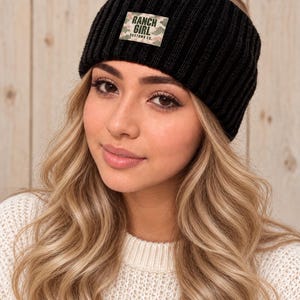 May include: A black ribbed knit headband featuring a rectangular patch on the front. The patch has a camouflage pattern in shades of green, pink, and white, with the text RANCH GIRL CUSTOMS CO. printed in black capital letters.