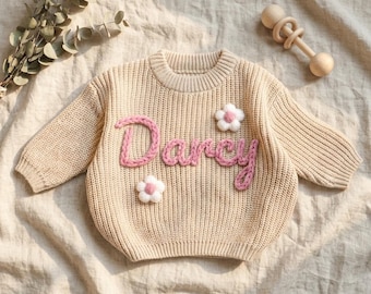 Op de afbeelding: Een beige gebreide trui met lange mouwen en de naam Darcy geborduurd in roze garen op de borst, versierd met twee kleine witte en roze bloemapplicaties, vergezeld van een houten rammelaar en een takje gedroogde eucalyptus.