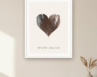 May include: A framed wall art print featuring a dark brown heart shape formed by two overlapping fingerprints on a cream background. Below the heart, the text reads Mr & Mrs · June 2026 in a dark serif font. The print is enclosed in a white frame.