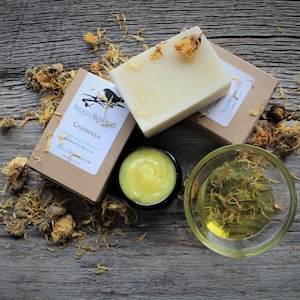 Op de afbeelding: Een rustieke opstelling van natuurlijke huidverzorgingsproducten, waaronder twee zeepblokken, een kleine pot crème en een kom olie, allemaal doordrenkt met calendula. De zeepblokken zijn licht van kleur en versierd met gedroogde calendulabloemen. Gepresenteerd op een verweerd houten oppervlak.