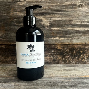 Op de afbeelding: Een zwarte pompflacon met pepermunt-theeboomolie handzeep van Indigo Sundries Soap Company. De fles is voorzien van een wit etiket en toont een kleine illustratie van twee vogels op een tak.