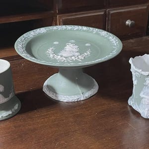 May include: A set of three sage green and white jasperware pieces including a cylindrical cup, a pedestal cake stand with a central floral relief, and a flared vase featuring a white relief of a person playing a stringed instrument.