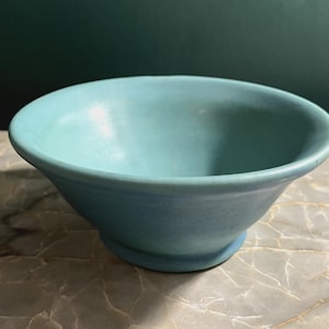 May include: A matte light blue ceramic bowl with a wide, flared rim and a circular base. The bowl features a smooth, solid-colored finish with subtle variations in tone across its surface, creating a simple and functional design.