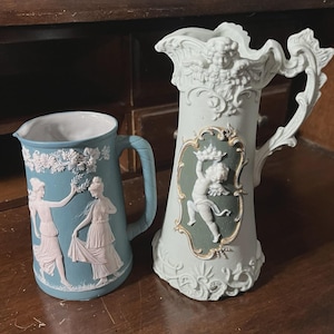 May include: A light blue ceramic pitcher with white relief figures of two women and a taller, pale green ceramic pitcher featuring a white relief of a cherub holding a fruit basket, both with ornate handles and detailed decorative borders.