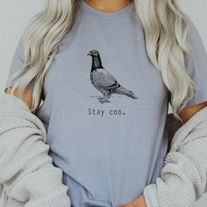 以下が含まれることがあります： 薄い紫色のTシャツに、白黒の鳩のイラストと「Stay coo.」というテキストがプリントされています