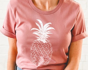Pode incluir: Uma camiseta rosa com um gráfico de abacaxi branco. O abacaxi é um design simples e estilizado com um visual texturizado.