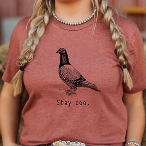 以下が含まれることがあります： さび色のTシャツに、白黒の鳩のイラストと「Stay coo.」の文字がプリントされています