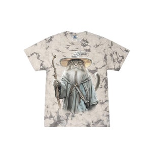May include: A tie-dye t-shirt with a light brown background features a cat dressed as a wizard. The cat is wearing a pointed hat, a long blue robe, and holding a staff. The cat has a long white beard and a serious expression.