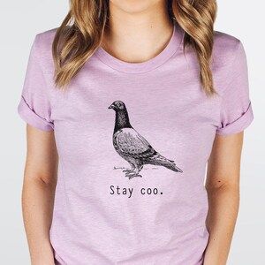 以下が含まれることがあります： 薄い紫色のTシャツに、白黒の鳩のイラストと「Stay coo.」という文字がプリントされています