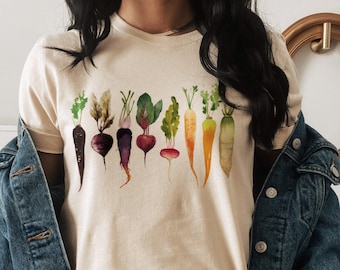 May include: A beige t-shirt featuring a watercolour illustration of a row of colourful vegetables, including carrots, radishes, and beetroot.