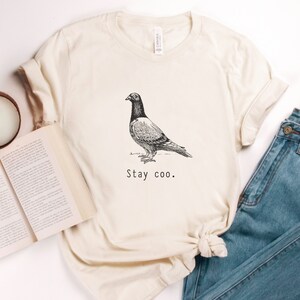 以下が含まれることがあります： クリーム色のTシャツに、白黒の鳩のイラストと「Stay coo.」の文字がプリントされています