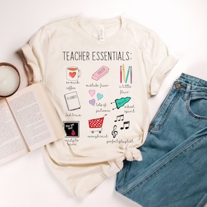 May include: A beige t-shirt with the text "TEACHER ESSENTIALS:" and a list of essential items for teachers, including coffee, a planner, a red eraser, pencils, hearts, a shopping cart, music notes, and a chalkboard with the text "Now you see it".
