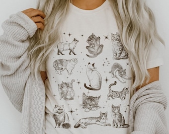 May include: A white t-shirt with a black and white illustration of various cats. The cats are in different poses, some are sitting, some are standing, and some are lying down. The shirt has a crew neck and short sleeves.