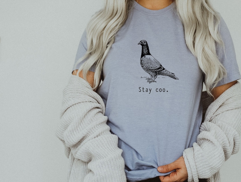 以下が含まれることがあります： 薄い紫色のTシャツに、白黒の鳩のイラストと「Stay coo.」というテキストがプリントされています