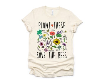 May include: A beige t-shirt featuring a graphic of flowers and bees. The text "PLANT THESE" and "SAVE THE BEES" is printed on the shirt. The flowers include poppy, sunflower, lavender, wild rose, sage, and forget-me-not.