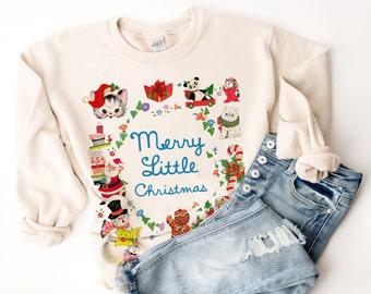 Puede incluir: Una sudadera de color crema con un diseño navideño que presenta una corona de acebo, bastones de caramelo y varios animales. El texto "Merry Little Christmas" está impreso en azul en la parte delantera de la sudadera.