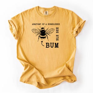 Puede incluir: Camiseta de color amarillo mostaza con un gráfico de abejorro y el texto "ANATOMY OF A BUMBLEBEE" y "BLE BEE BUM" en negro. La camiseta tiene mangas cortas y cuello redondo.