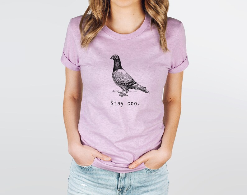 以下が含まれることがあります： 薄い紫色のTシャツに、白黒の鳩のイラストと「Stay coo.」という文字がプリントされています