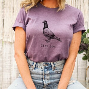 以下が含まれることがあります： 鳩の白黒のイラストと「Stay coo.」の文字が入った紫色のTシャツ