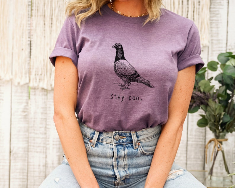 以下が含まれることがあります： 鳩の白黒のイラストと「Stay coo.」の文字が入った紫色のTシャツ