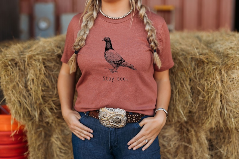 以下が含まれることがあります： さび色のTシャツに、白黒の鳩のイラストと「Stay coo.」の文字がプリントされています