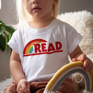 May include: White t-shirt with a rainbow graphic and the word "READ" in red. The shirt is worn by a child. The rainbow design features red, orange, yellow, green, and blue colors. The shirt is made of a soft material.