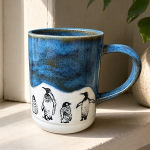 May include: A ceramic mug featuring a two-tone design with a deep blue glazed upper section and a white lower section. The white base is decorated with four black ink-style illustrations of penguins in various poses, including standing and walking.