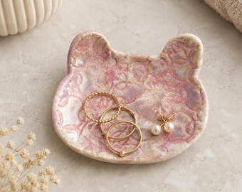 May include: A small, cat-shaped ceramic trinket dish featuring a mottled pink and lavender glaze with an embossed floral pattern. The dish holds two gold-toned rings and a pair of small gold earrings with white pearl-like accents.