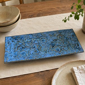 May include: A rectangular ceramic serving platter featuring a vibrant blue glaze with a repeating embossed heart pattern and dark, weathered detailing along the edges and recessed areas, showcasing a textured, hand-crafted appearance.